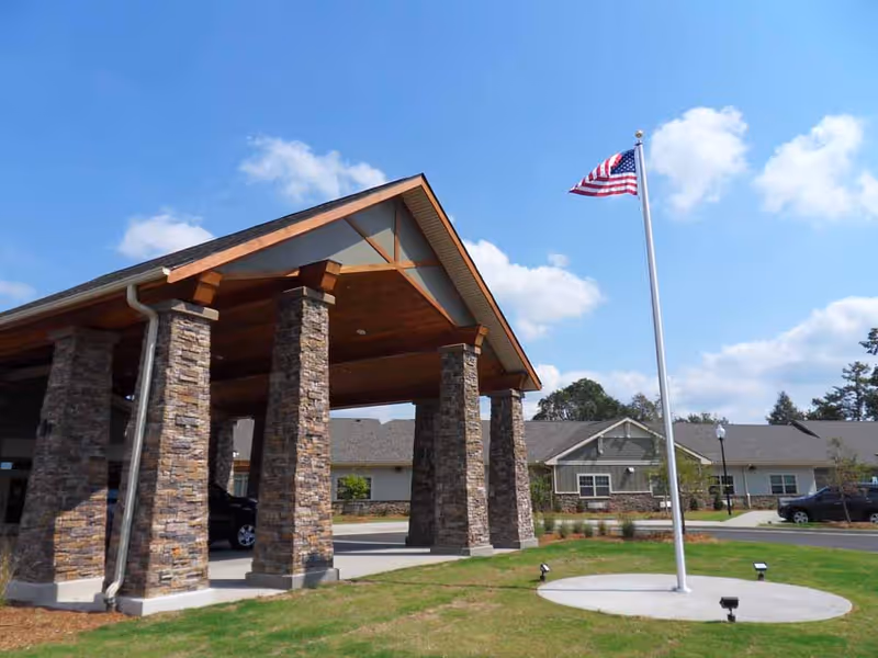 Front entrance of a senior living building with a stone-pillared covered drive and an American flag on a flagpole.
