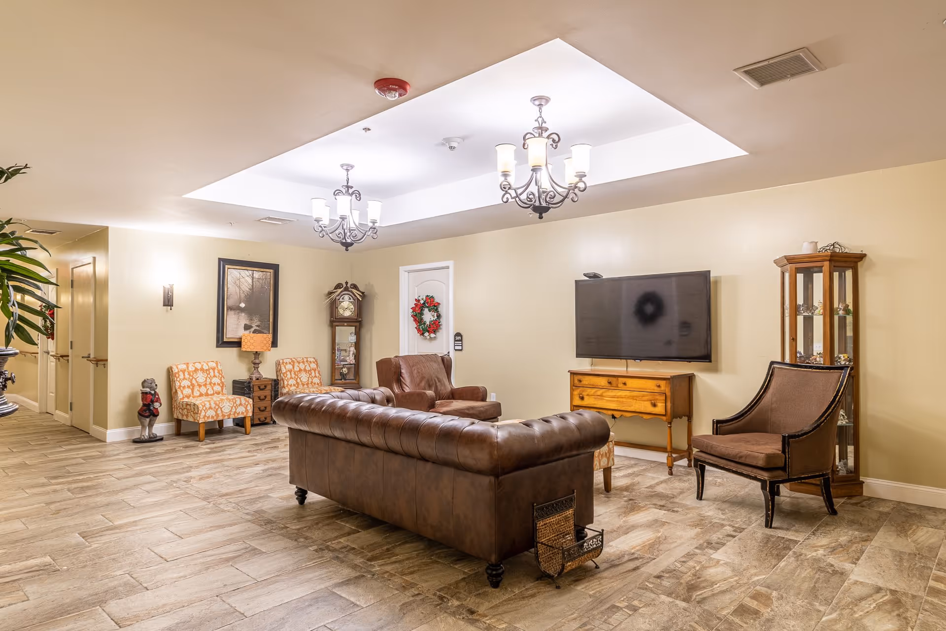 Spacious lounge with a brown leather sofa, armchairs, a wall-mounted TV, chandeliers, and decorative furniture.