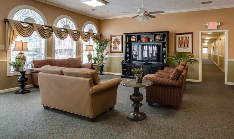 A cozy living room area in a senior living facility with three upholstered sofas arranged around a small round wooden table. The room features large arched windows with decorative valances, two table lamps on side tables, a black entertainment center with a TV, framed artwork on the walls, and potted plants. A hallway with doors and an exit sign is visible in the background.