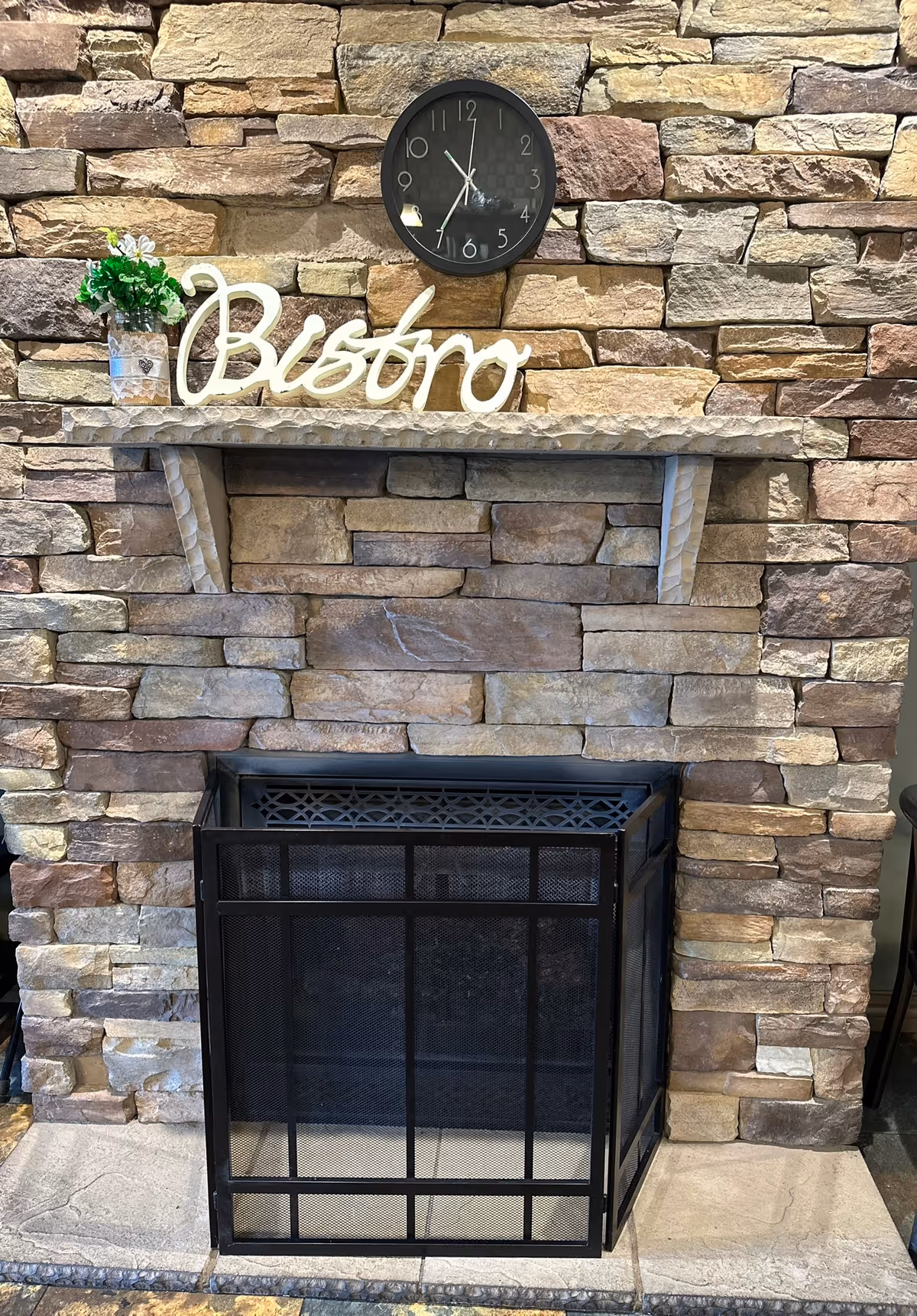 Stone fireplace with a black metal screen in front. Above the fireplace is a stone mantel holding a small potted plant and a decorative sign that reads 'Bistro'. A round black clock is mounted on the stone wall above the mantel.