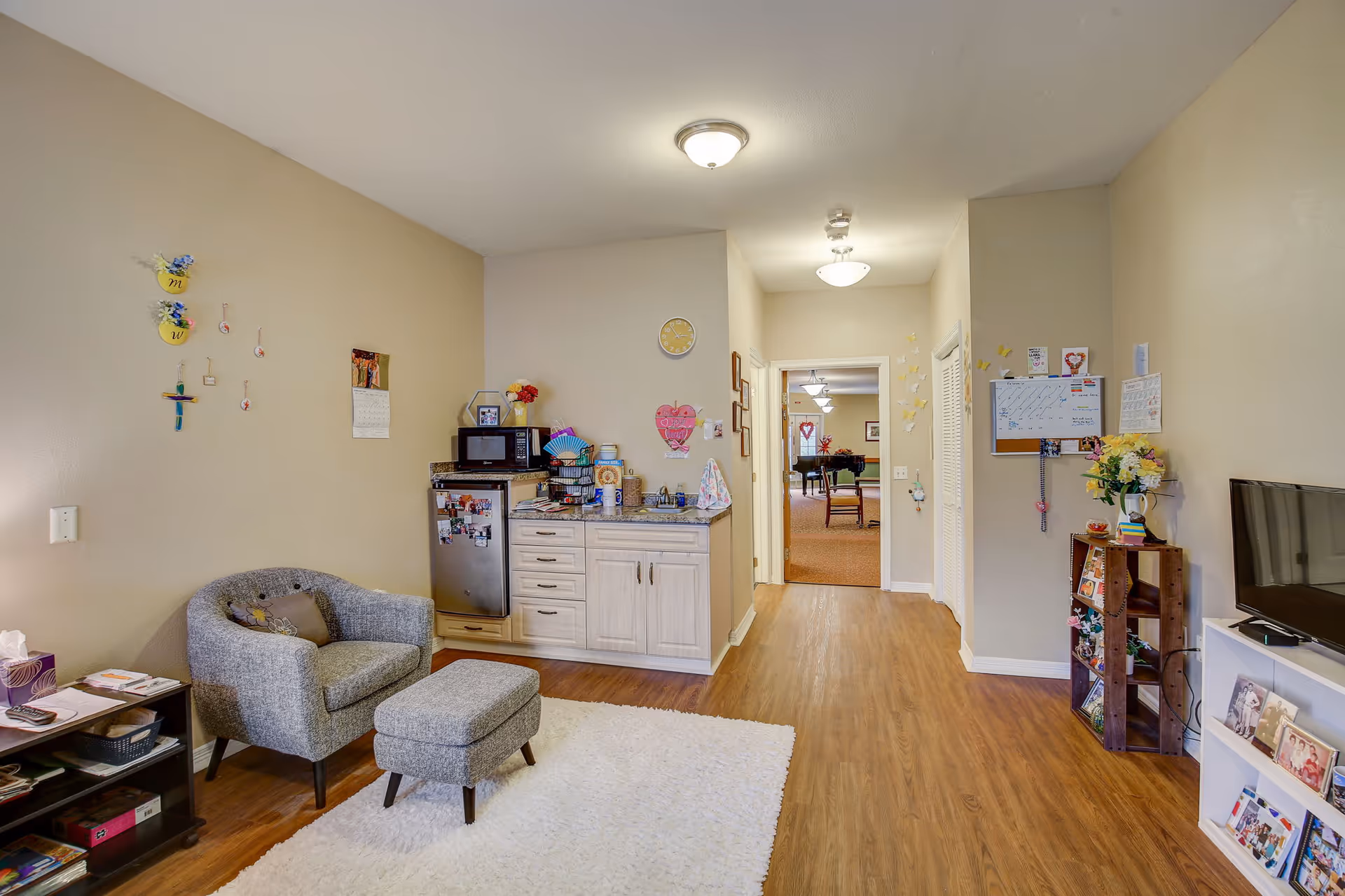 Comfortable living room area with a gray armchair and ottoman, small kitchenette/cabinetry, and a TV in a senior living facility.