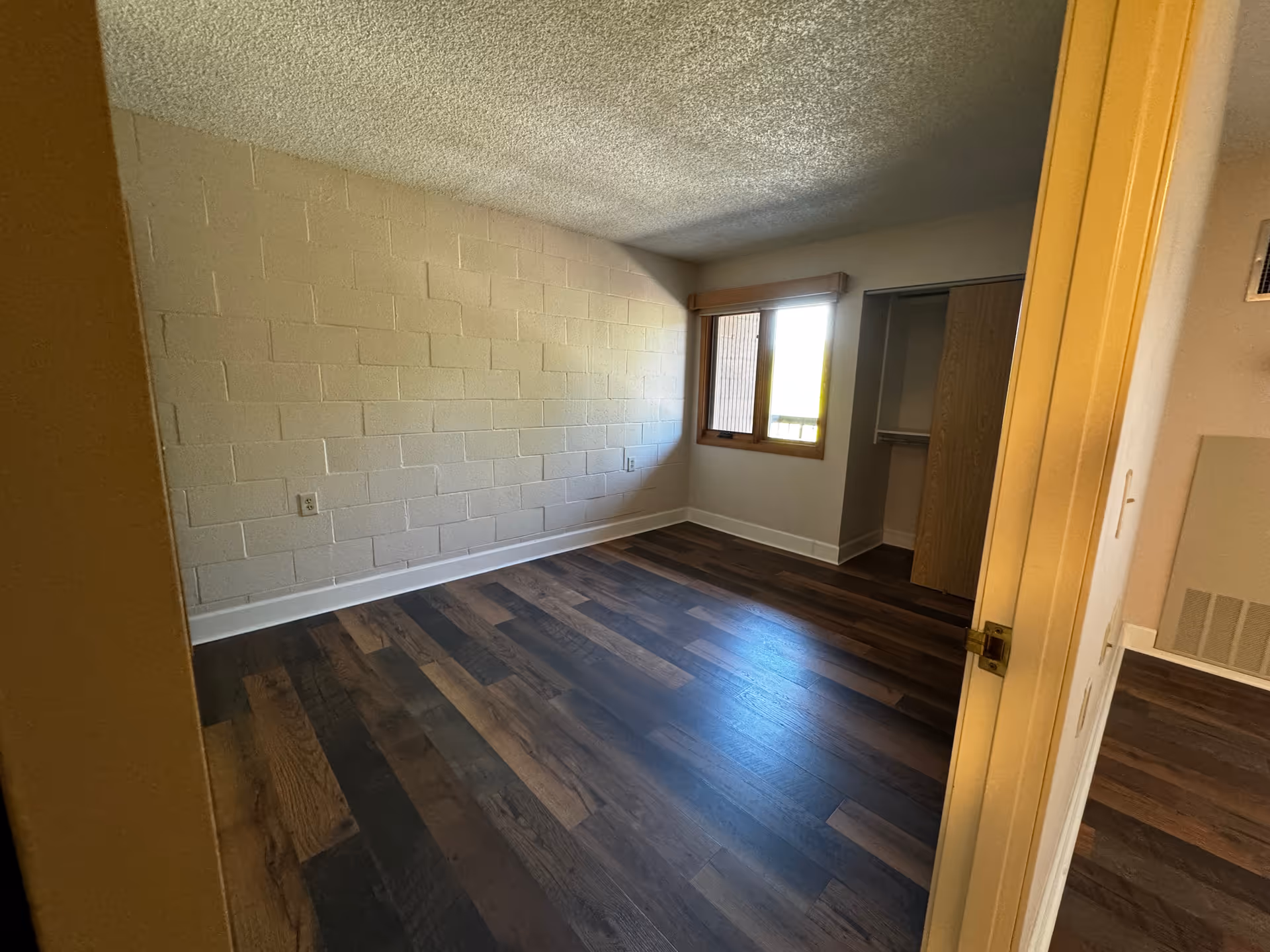Empty bedroom with dark wood-look floors, a cinderblock accent wall, a window with blinds and an open closet seen from the doorway.