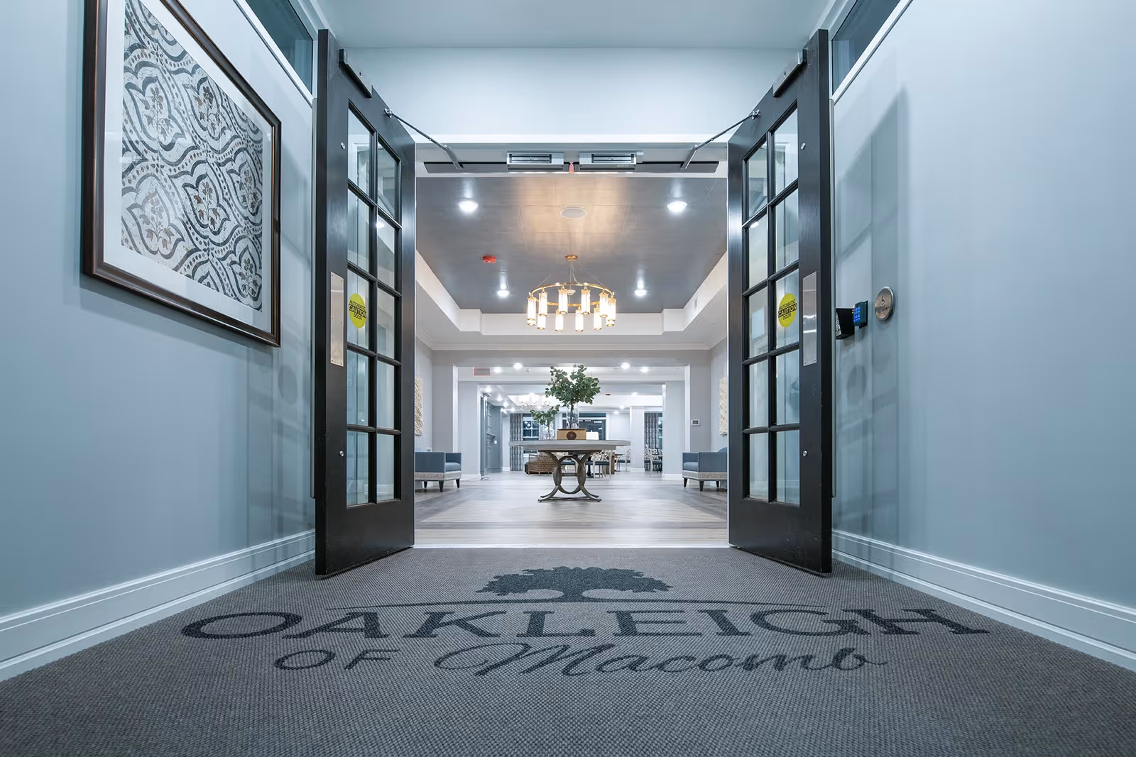Entrance hallway of Oakleigh of Macomb Senior Living with double glass doors opening into a spacious, well-lit common area featuring a central table with a plant and seating along the walls.