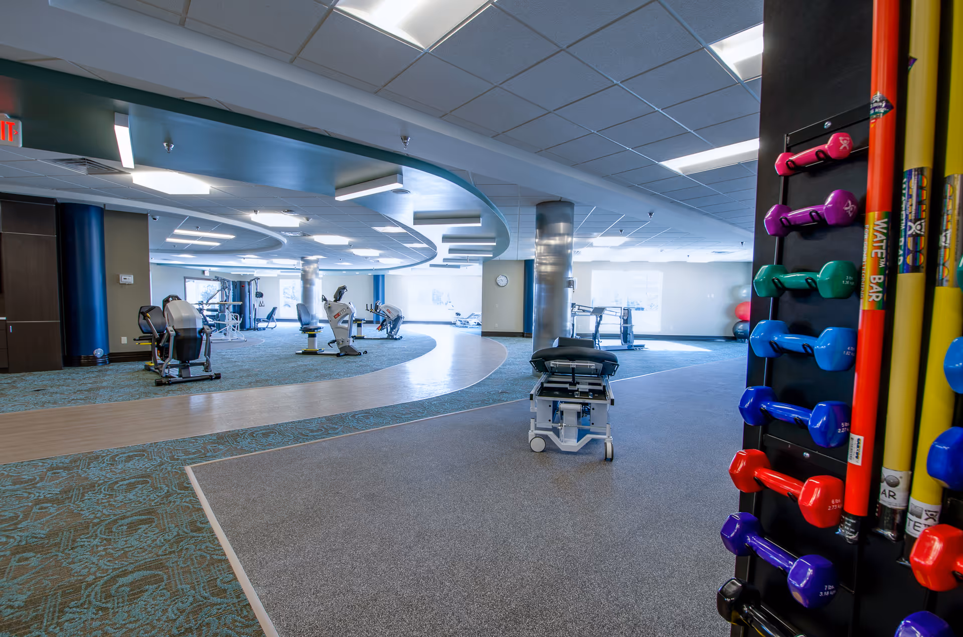 A spacious rehabilitation gym area with exercise equipment including stationary bikes and weight racks. The room has a modern design with a curved ceiling feature, large windows letting in natural light, and a variety of colorful dumbbells and exercise bars on a rack in the foreground.