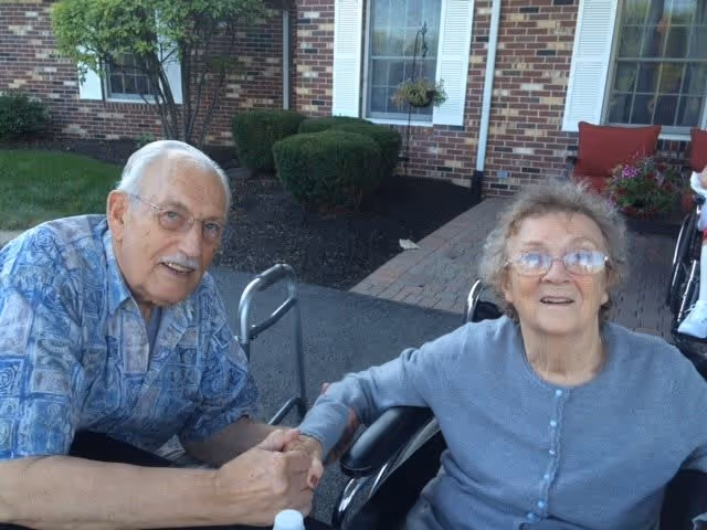 An elderly man and woman sitting outside near a brick building with windows and white shutters. The man is wearing glasses and a blue patterned shirt, holding the woman's hand. The woman is wearing glasses and a light blue cardigan, sitting in a wheelchair. There are bushes and a small tree in the background.