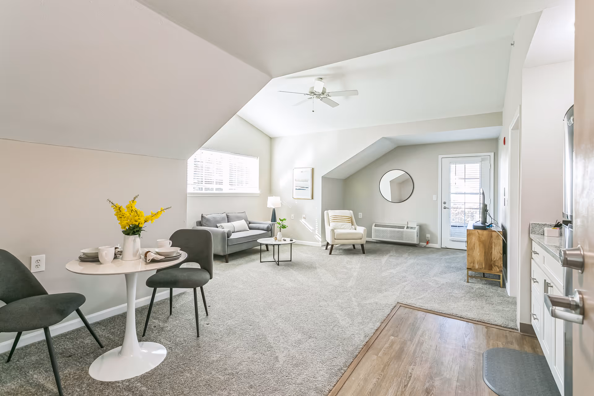 Bright open living room with a small round dining table, sofa, armchair, and kitchenette under a vaulted ceiling.