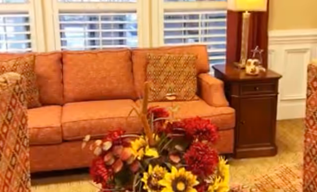 A cozy living room area featuring an orange patterned sofa with matching cushions, a wooden side table with a lamp and decorative items, and a floral arrangement with red and yellow flowers in the foreground. White window blinds and beige walls with white wainscoting are visible in the background.