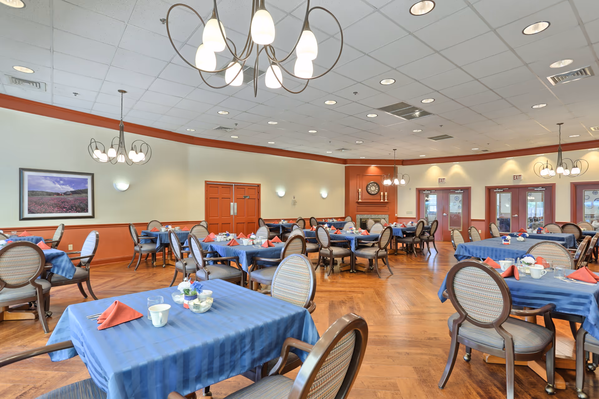 Spacious dining room with multiple round and rectangular tables set with blue tablecloths and red napkins, chandeliers, and a fireplace.