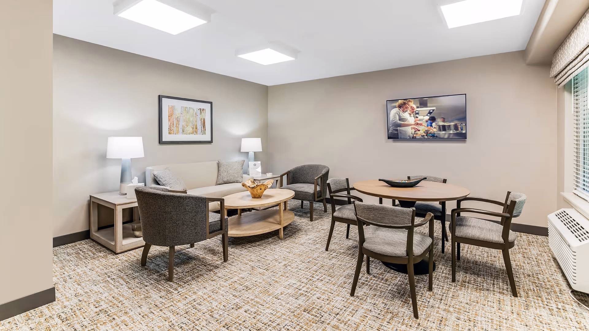 A cozy living room area in a senior living facility with a beige sofa, two gray armchairs, a wooden coffee table, and a round dining table with four chairs. The room has neutral-colored walls, carpeted flooring, two table lamps on side tables, a framed artwork on the wall, and a flat-screen TV mounted on the wall showing a cooking scene. A window with blinds and a heating unit are visible on the right side.