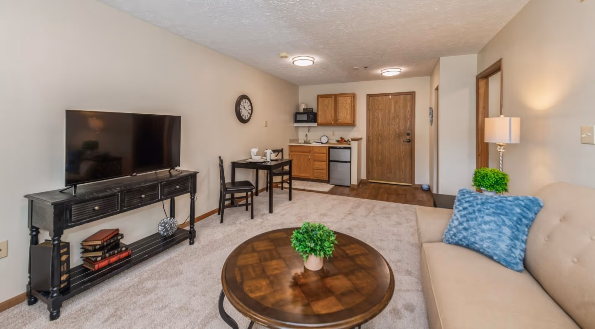 Open living area with a sofa and blue pillow, round coffee table, TV on a console, and a kitchenette and entrance door in the background.