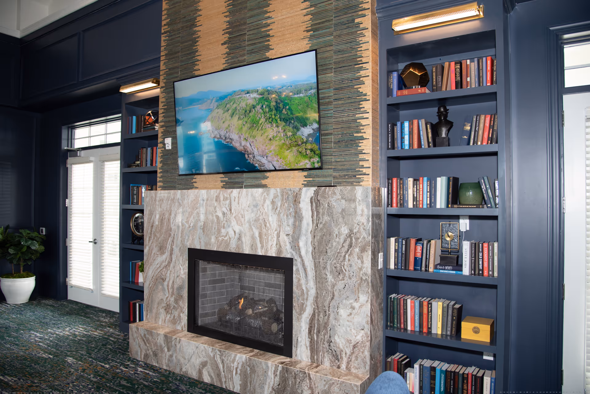 A cozy interior room with a marble fireplace and a flat-screen TV mounted above it displaying a scenic aerial view of a coastal landscape. On either side of the fireplace are built-in dark blue bookshelves filled with books and decorative items. There are double glass doors with white blinds on the left and right sides, and a potted plant is visible near the left door.