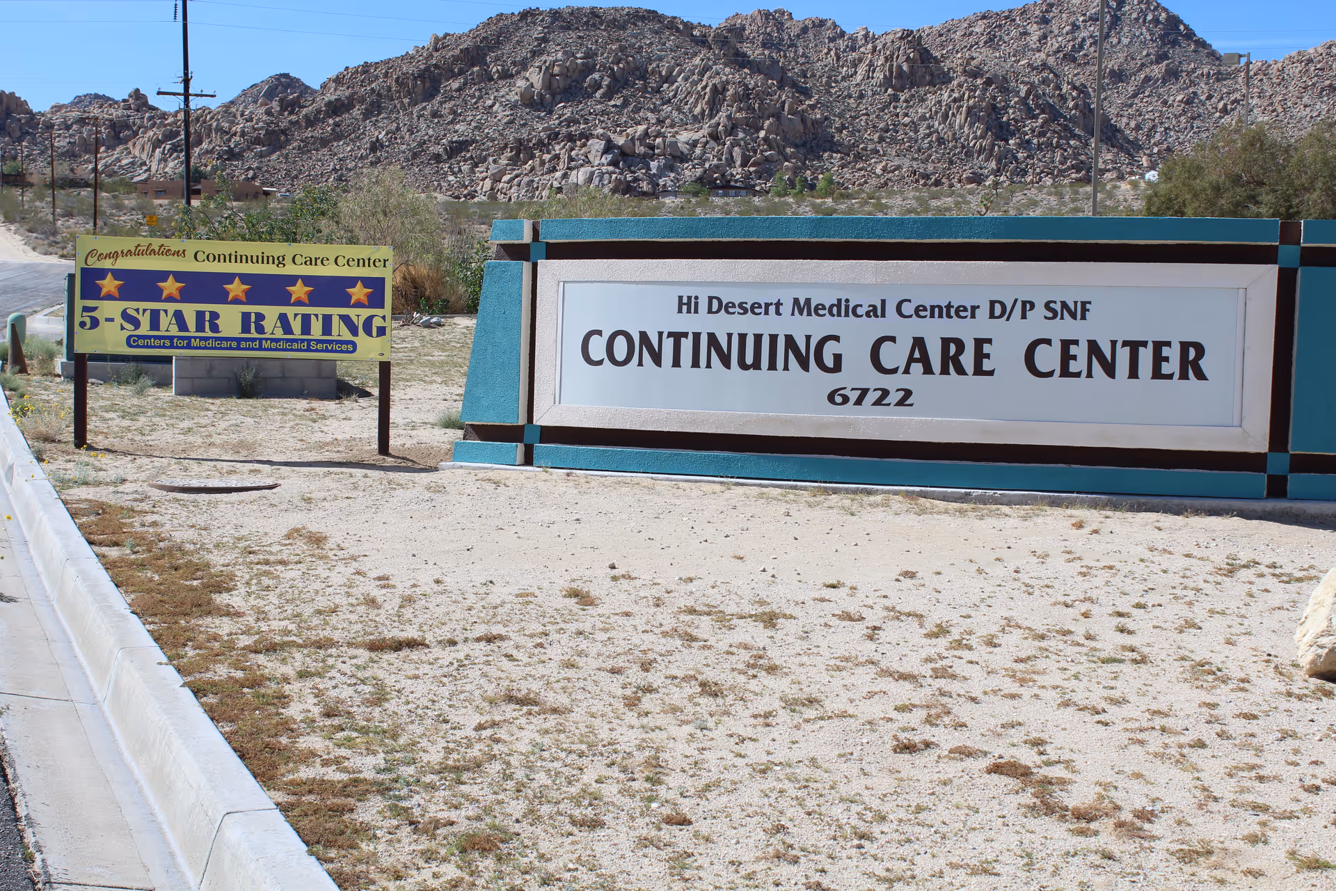 Outdoor view of the entrance signs for Hi Desert Medical Center Continuing Care Center, with a rocky hill in the background. One sign congratulates the center for a 5-star rating from Centers for Medicare and Medicaid Services.