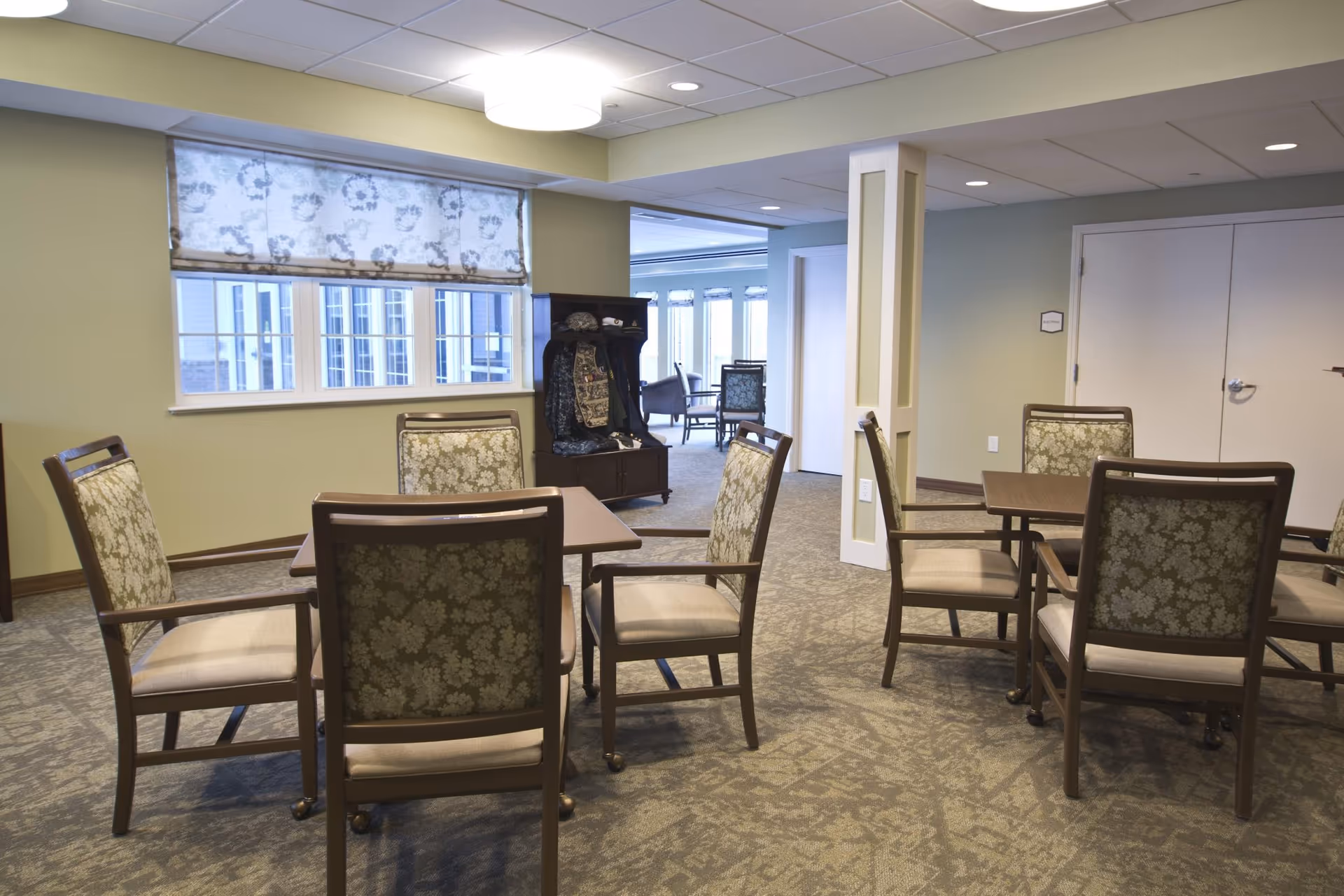 A well-lit interior room with several wooden tables and cushioned chairs arranged for seating. The room has light green walls, a window with a floral-patterned roller shade, and a carpeted floor. In the background, there is an open doorway leading to another room with more seating and large windows letting in natural light.