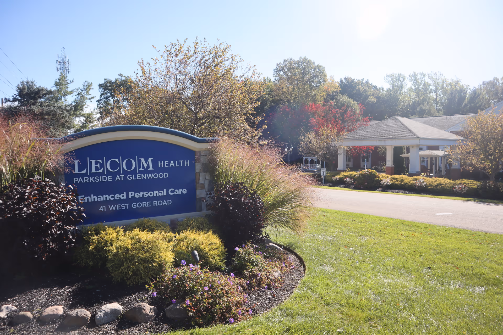 Front entrance and landscaped lawn of LECOM Health Parkside at Glenwood with a blue sign reading 'Enhanced Personal Care' and the building portico visible.