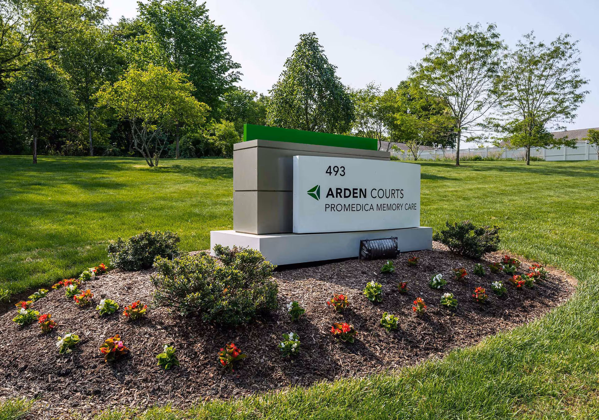 Landscaped lawn and flowerbed featuring a modern stone sign that reads 'Arden Courts ProMedica Memory Care' with trees in the background.