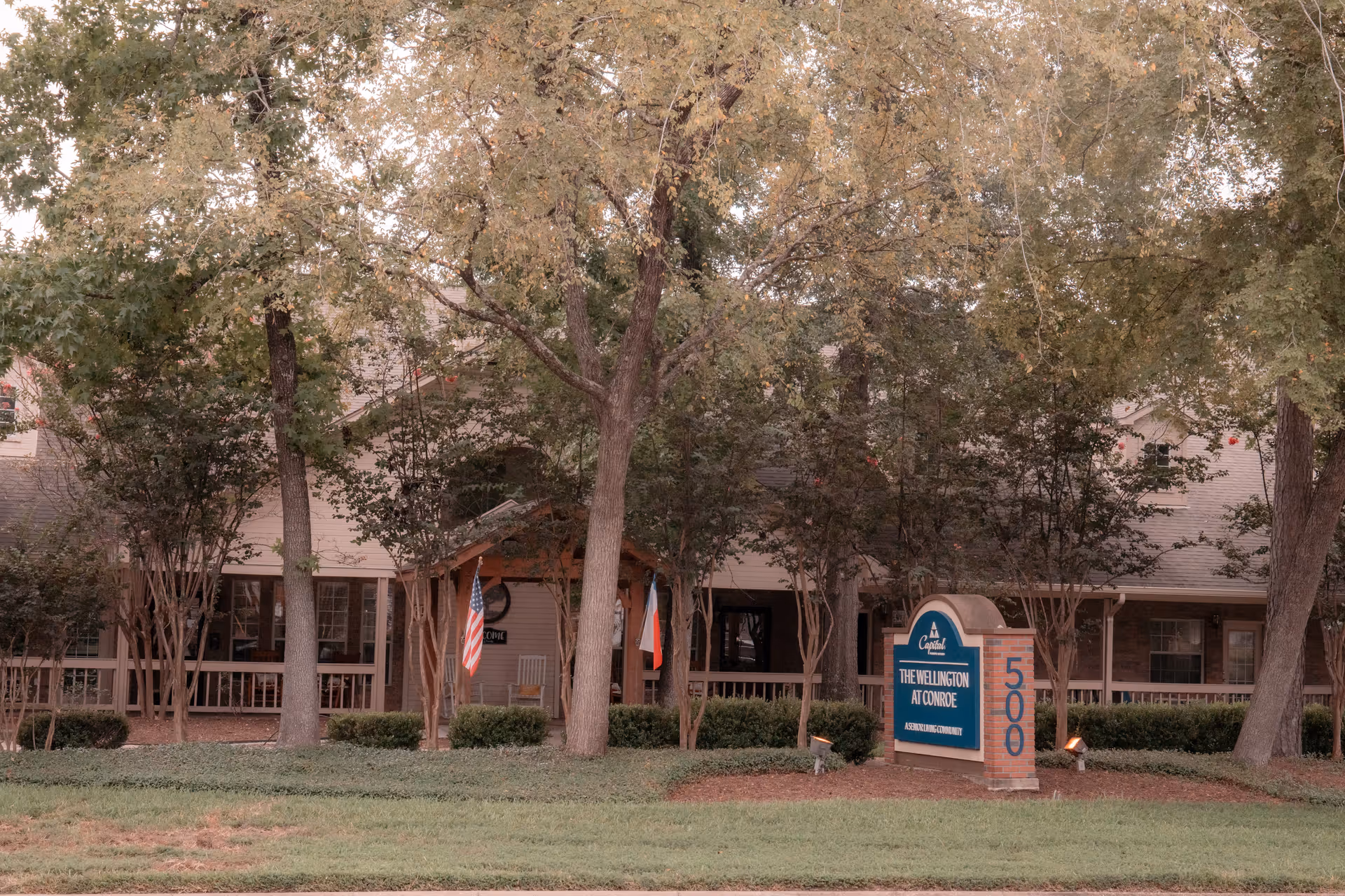 Exterior view of The Wellington at Conroe senior living community building partially obscured by trees and bushes, with a sign in front displaying the facility name and address number 500. Two flags are visible near the entrance porch.