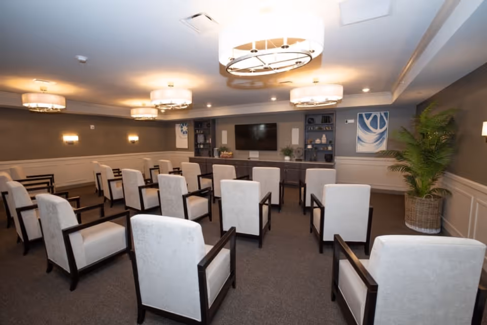 A meeting or presentation room with multiple beige upholstered chairs arranged in rows facing a large flat-screen TV mounted on a wall with built-in shelves. The room has gray walls, carpeted floor, modern ceiling lights, and a large potted plant in the corner.