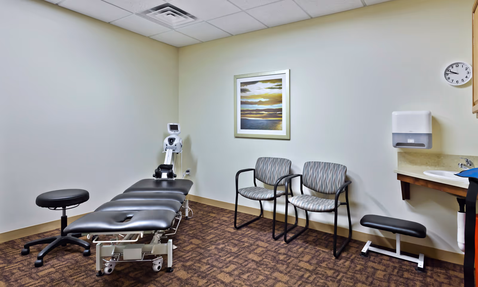 A clinical treatment room featuring an adjustable black therapy table, stools, two guest chairs, a sink, and framed artwork on the wall.