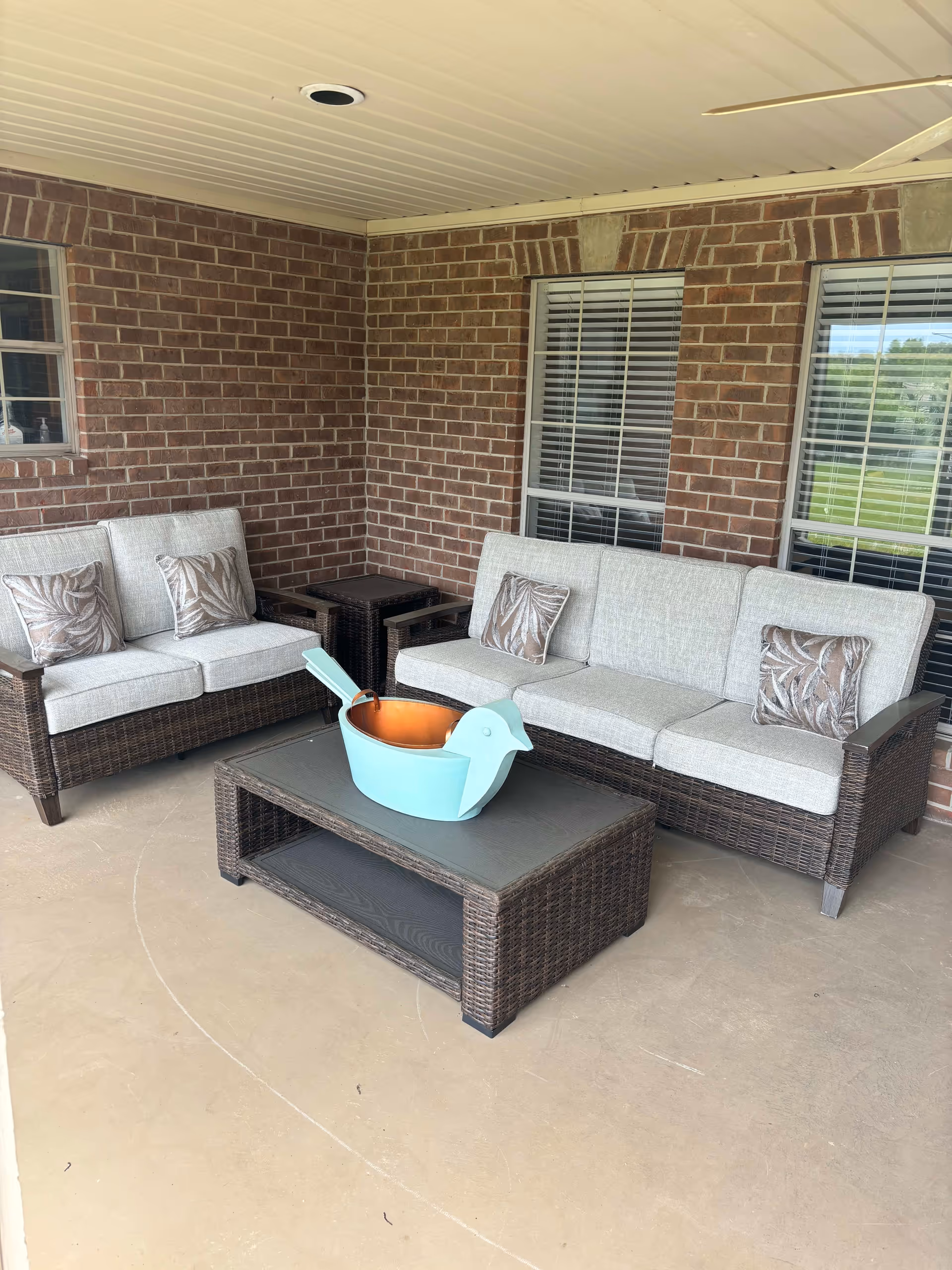 Covered outdoor patio area with two wicker sofas with light gray cushions and patterned pillows, a wicker coffee table with a large decorative blue bird-shaped container, and two windows with white blinds on a brick wall.