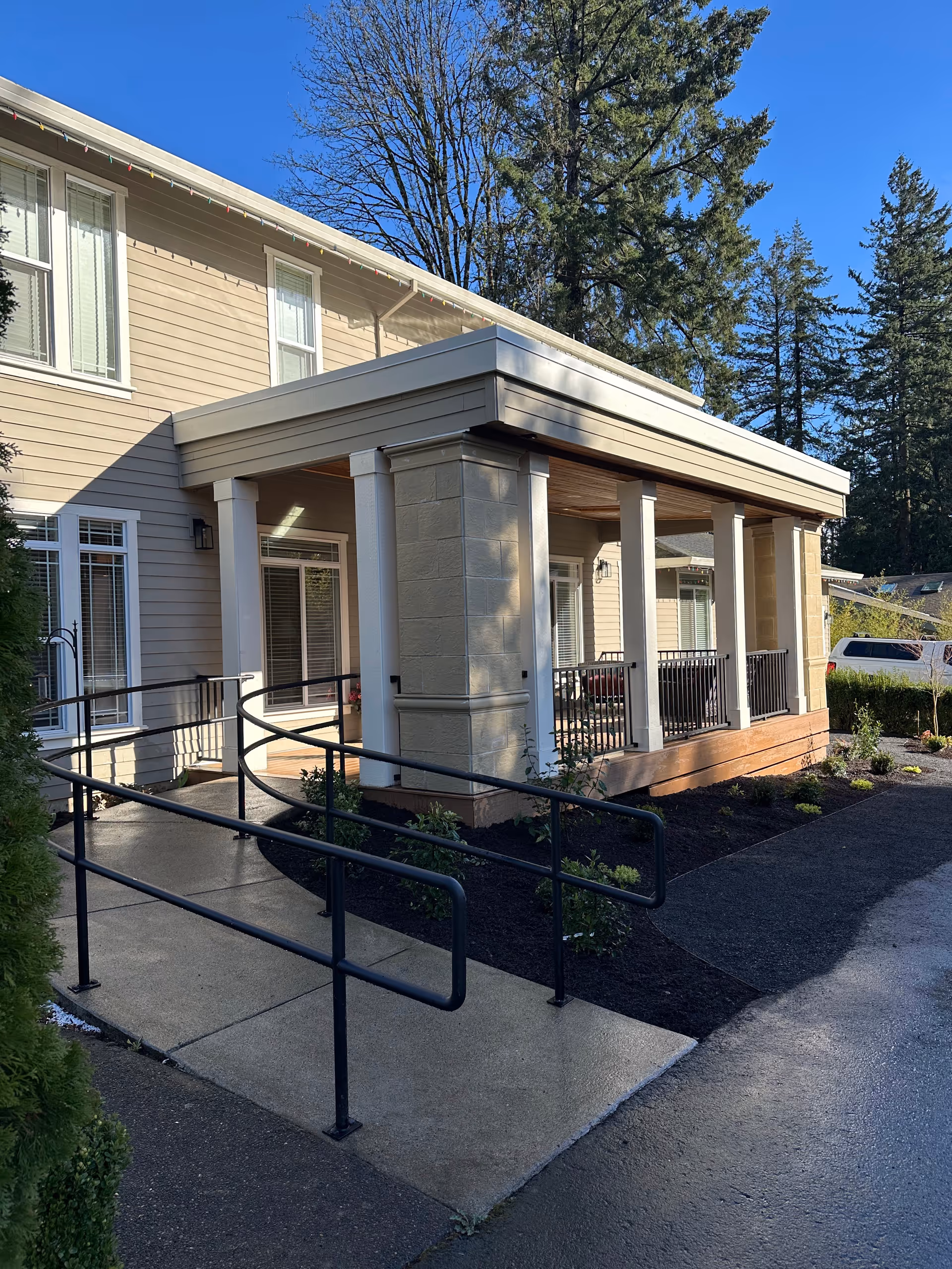 Entrance of a two-story beige building with a covered porch, columns, a wheelchair ramp, and surrounding landscaping and trees.