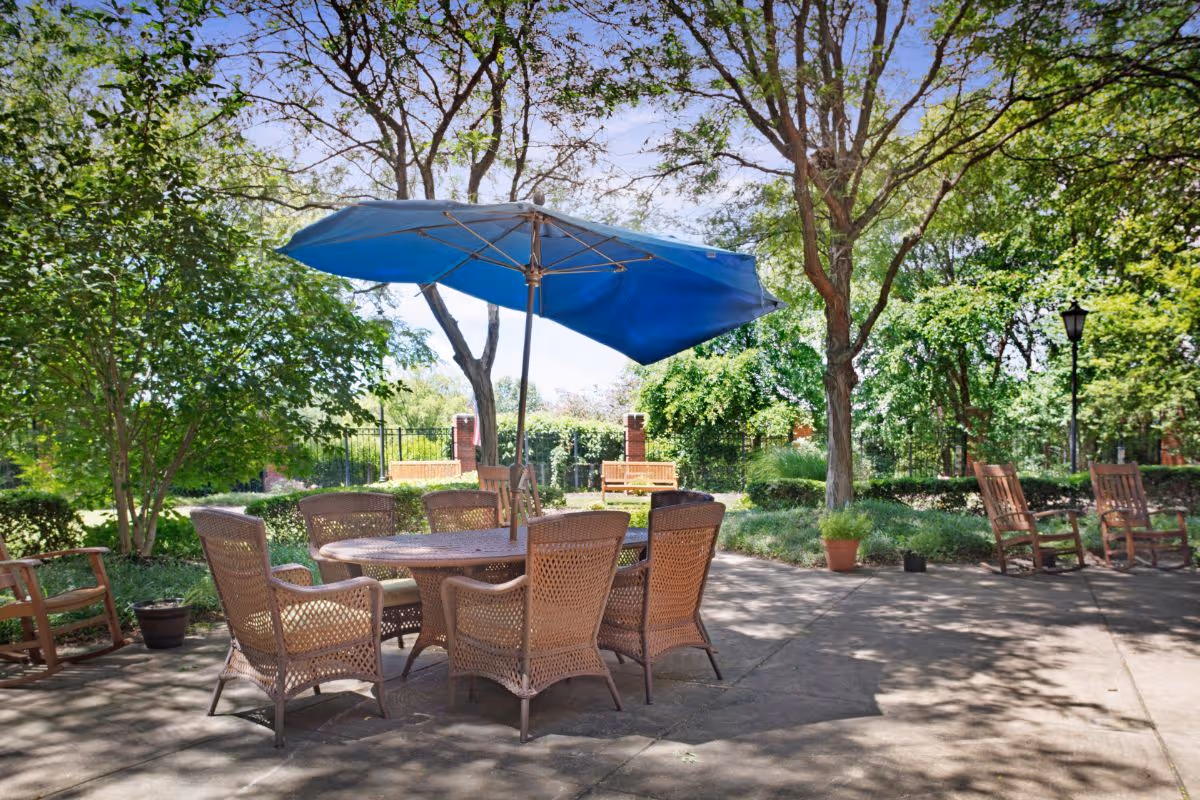 Outdoor patio area with a round table surrounded by six wicker chairs under a large blue umbrella. The patio is shaded by trees and has additional wooden rocking chairs and benches nearby, with greenery and a clear sky in the background.