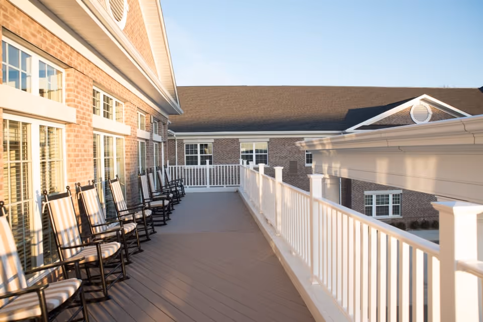 Outdoor balcony area at Harrison Trail Health Campus with a row of black rocking chairs with striped cushions lined up against a brick wall with large windows. The balcony has white railings and overlooks part of the building under a clear blue sky.