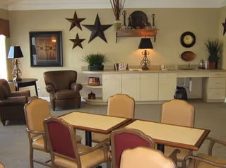 A cozy common area with a table and several chairs in the foreground. In the background, there are two brown armchairs, a countertop with cabinets underneath, decorative stars on the wall, a framed picture, a lamp, and some plants and decorative items.