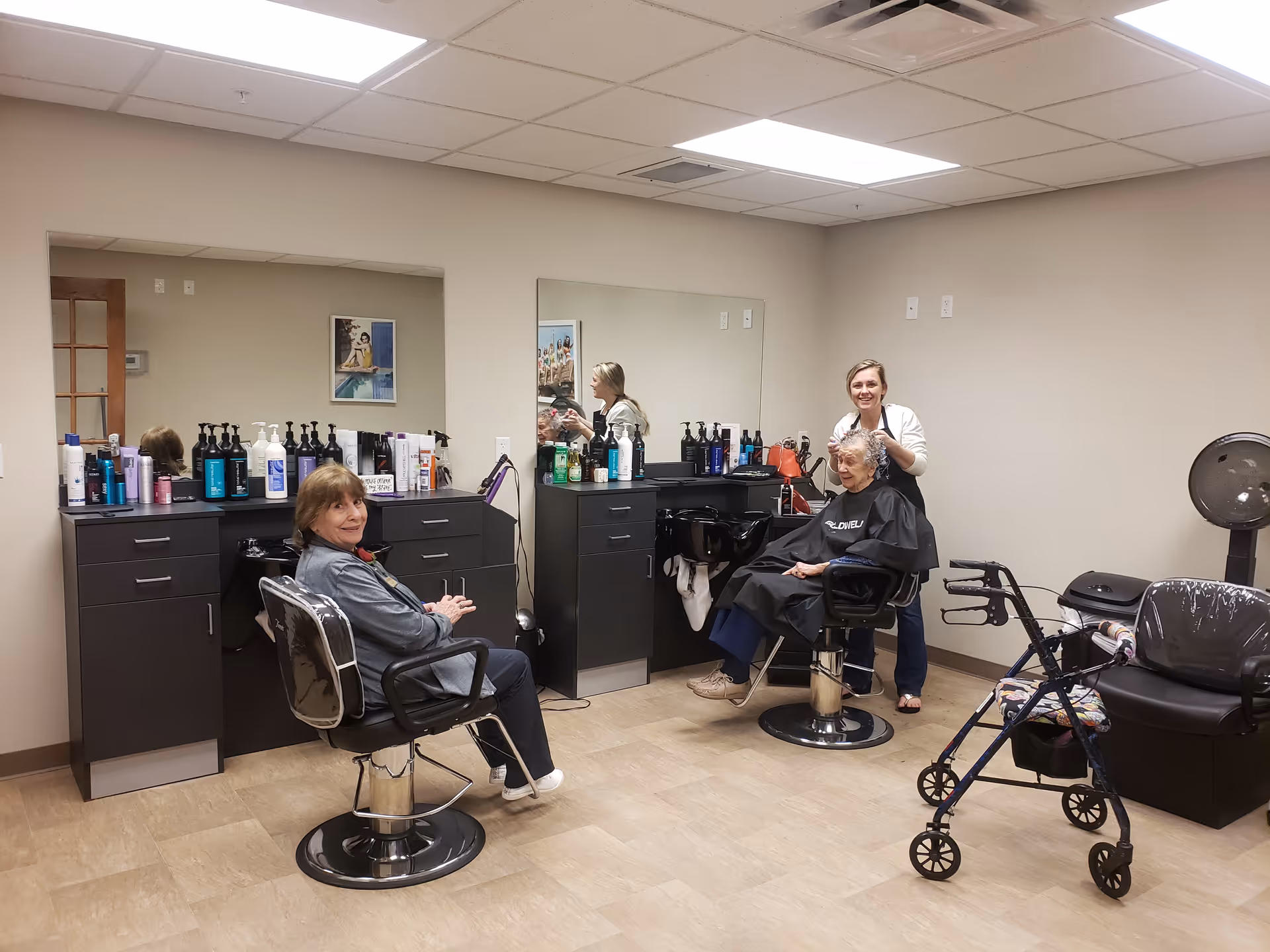 Interior of a hair salon at a retirement resort with two elderly women seated in salon chairs. One woman is sitting and smiling at the camera, while the other is having her hair styled by a hairdresser. The room has beige walls, a large mirror, black cabinets with various hair care products, and a walker nearby.