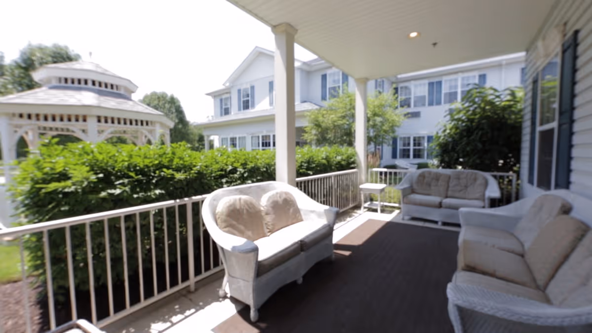 Covered outdoor patio area with cushioned wicker seating including two loveseats and a sofa, overlooking green bushes and a white gazebo with a multi-story building in the background.