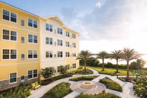 Yellow multi-story building beside a landscaped courtyard with a circular fountain, palm trees, and winding walkways at sunset.