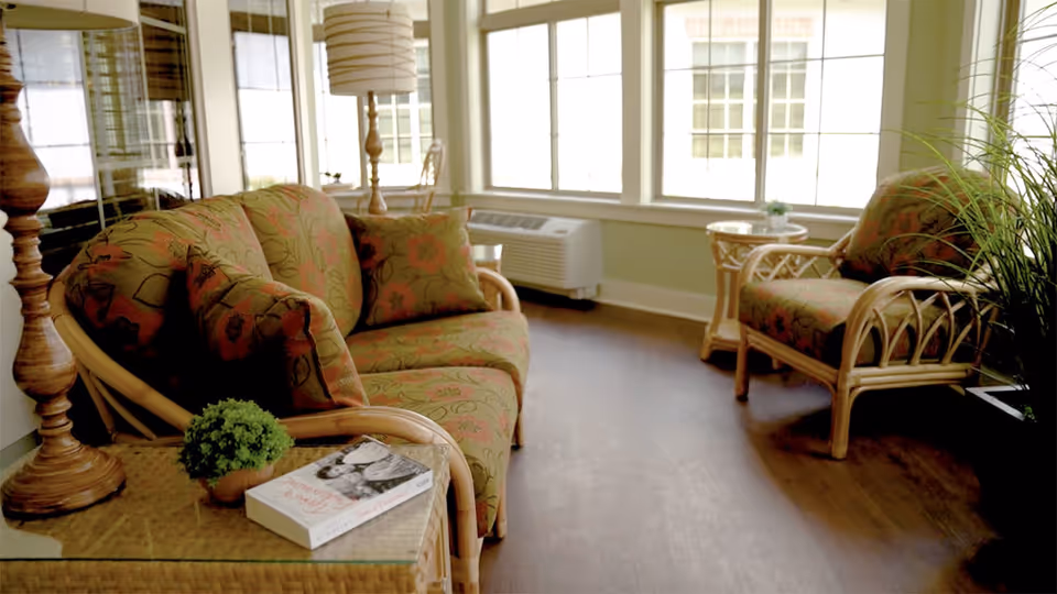 A cozy sitting area with a floral-patterned sofa and matching armchair made of light wood. There is a small glass-top side table with a potted plant and a book on it. The room has large windows letting in natural light, a floor lamp, and a wooden floor.