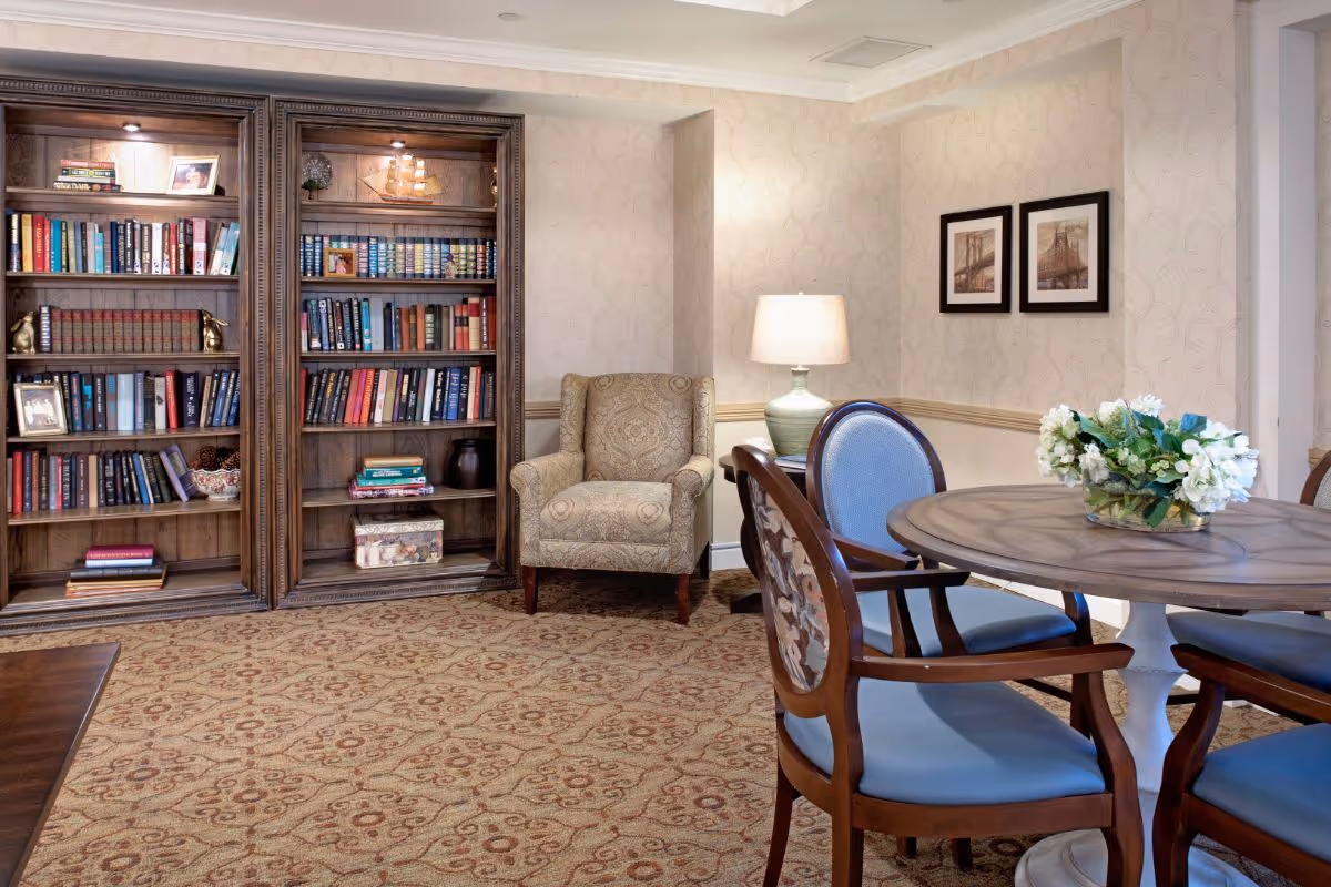 A cozy interior common room with wooden bookshelves, an upholstered armchair, a round table with chairs, and a table lamp.