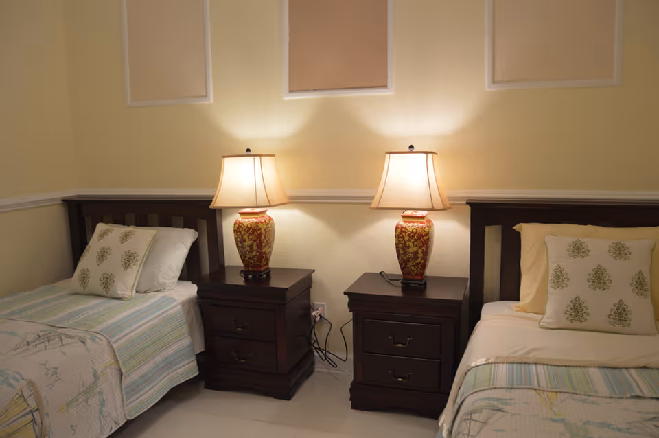 A bedroom with two single beds separated by two dark wooden nightstands, each with a decorative table lamp. The beds have light-colored bedding with patterned pillows. The walls are painted light yellow with three framed fabric panels above the beds.