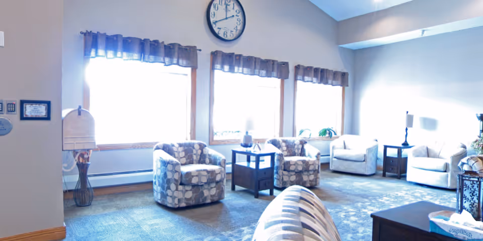 A bright living room area with multiple cushioned armchairs arranged around small wooden tables. There are three large windows with valance curtains letting in natural light, a wall clock above the windows, and a floor lamp in the corner. The room has carpeted flooring and light-colored walls.