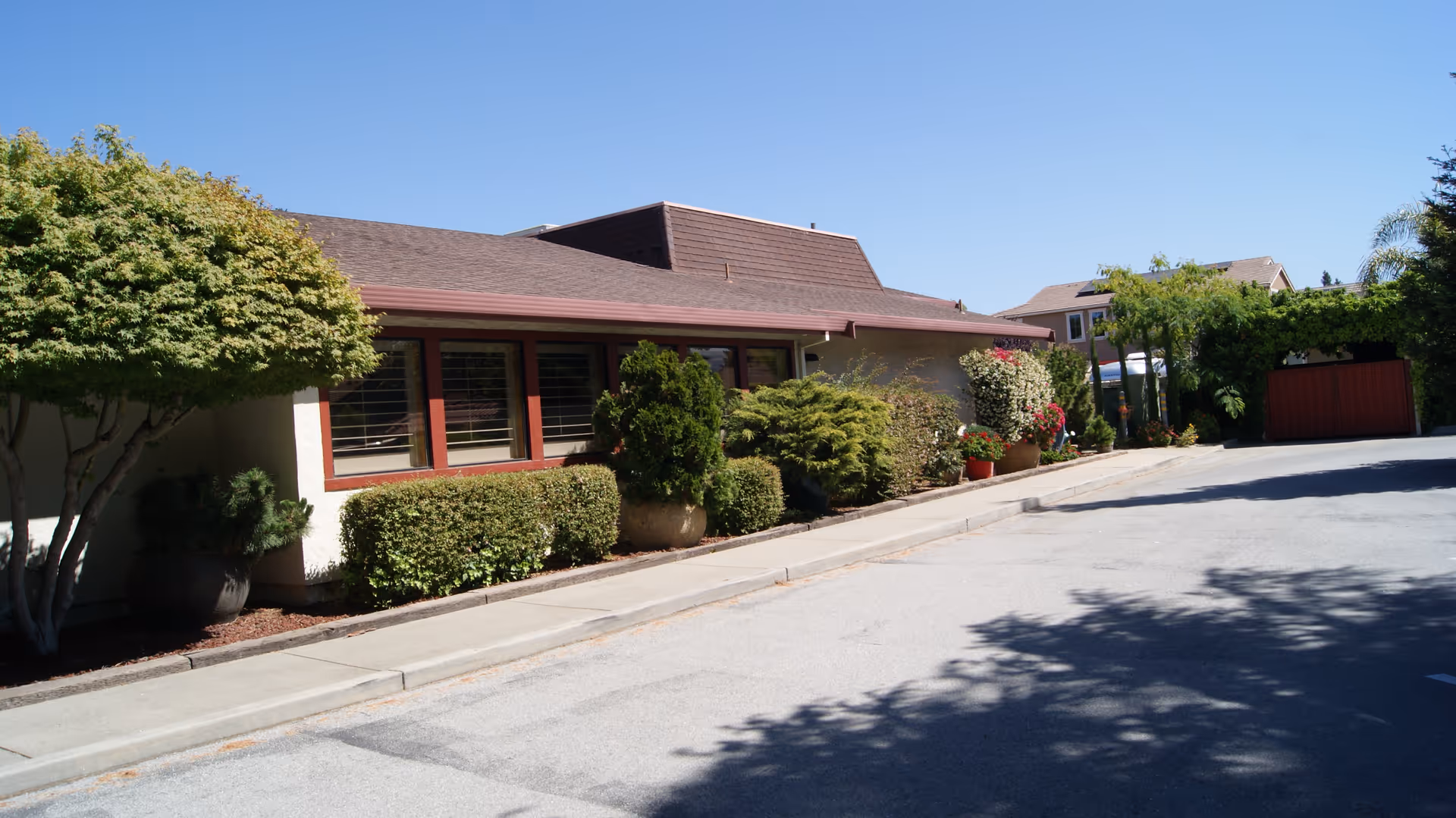 Exterior view of a single-story building with a sloped roof, surrounded by various green shrubs and potted plants along a sidewalk under a clear blue sky.