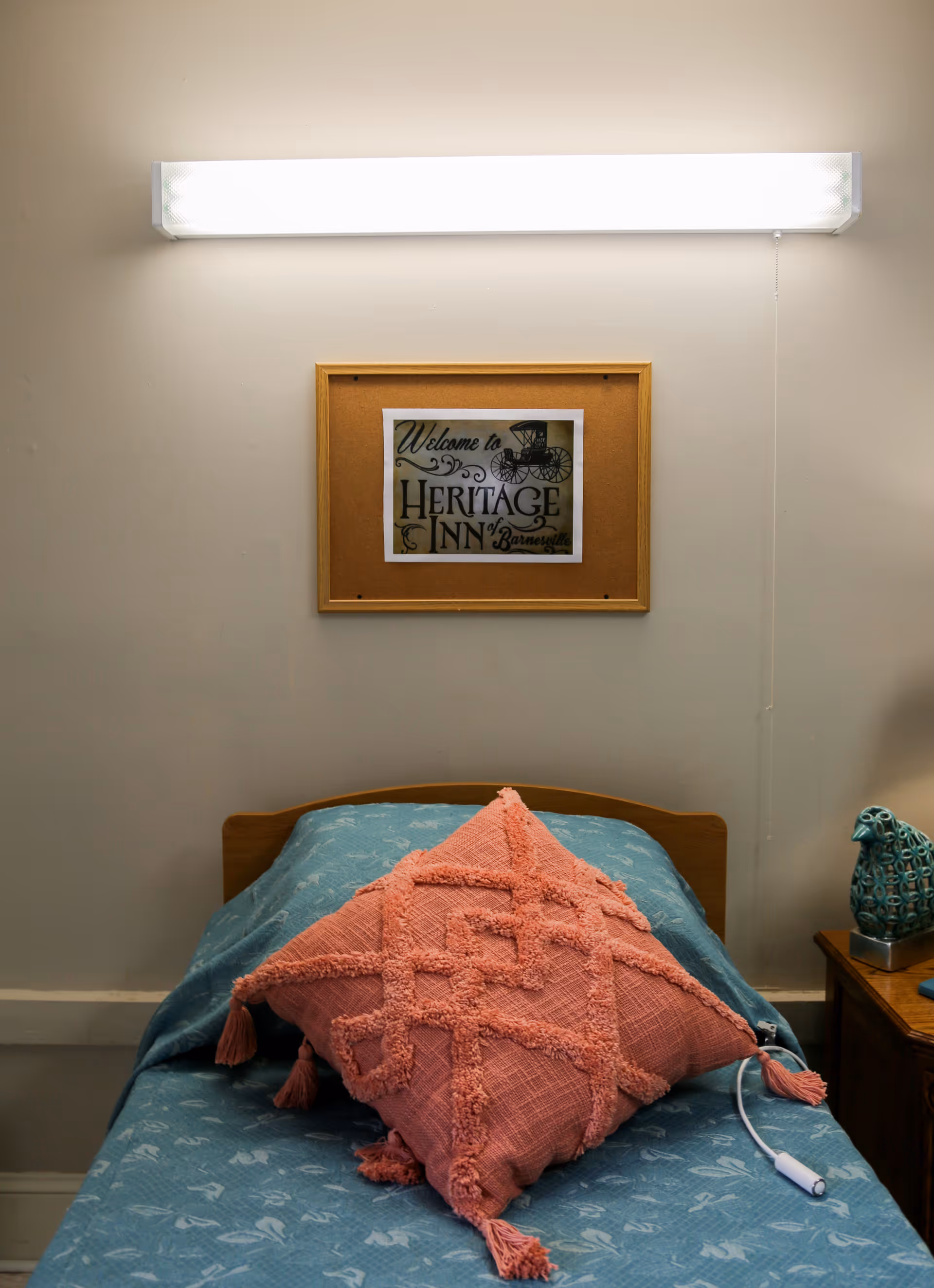 A single bed with a blue patterned bedspread and a large pink textured pillow placed on top. Above the bed is a framed sign that reads 'Welcome to Heritage Inn of Barnesville.' A bright fluorescent light fixture is mounted on the wall above the sign. To the right of the bed is a wooden nightstand with a decorative teal ceramic owl and a lamp.