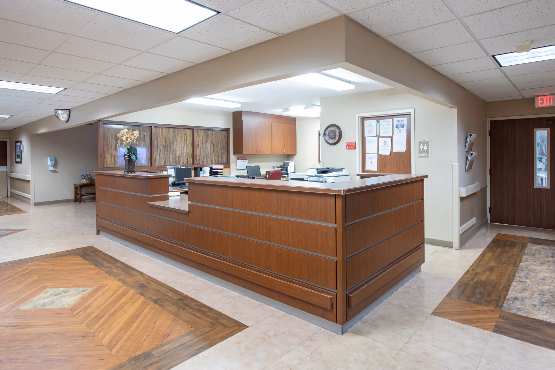 A clean and well-lit reception desk area inside a healthcare facility with wooden paneling, office equipment, and a flower vase on the counter. The surrounding area has tiled floors with wood-patterned inlays and beige walls with doors and signage.