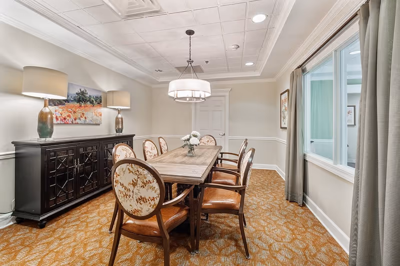 A well-lit dining room with a rectangular wooden table surrounded by six upholstered chairs with floral patterns. The room features a patterned carpet, a black sideboard with two table lamps, a floral painting on the wall, a modern chandelier hanging above the table, and a window with curtains on the right side.