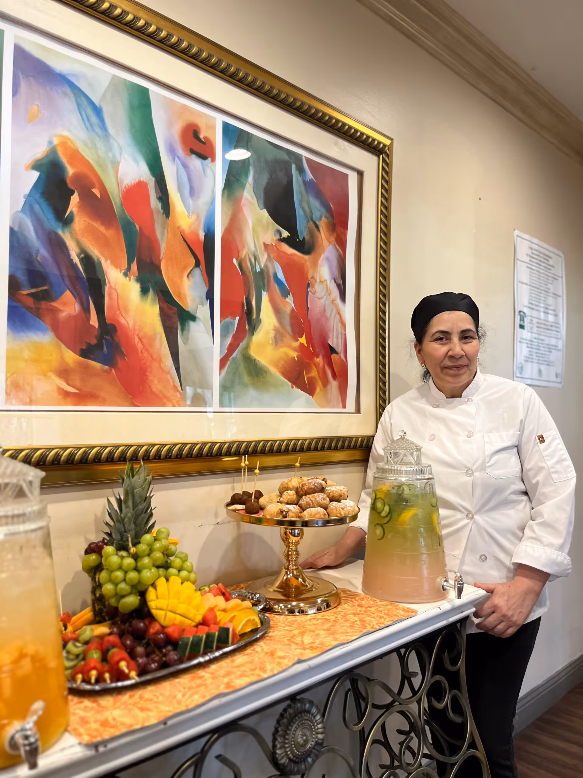 A woman in a white chef's coat and black head covering stands next to a table with a variety of food and drinks. The table has a large platter of fresh fruit including grapes, pineapple, mango, and watermelon, a tiered tray with pastries, and two large beverage dispensers with infused water and juice. Behind her is a wall with a large colorful abstract painting in a gold frame.