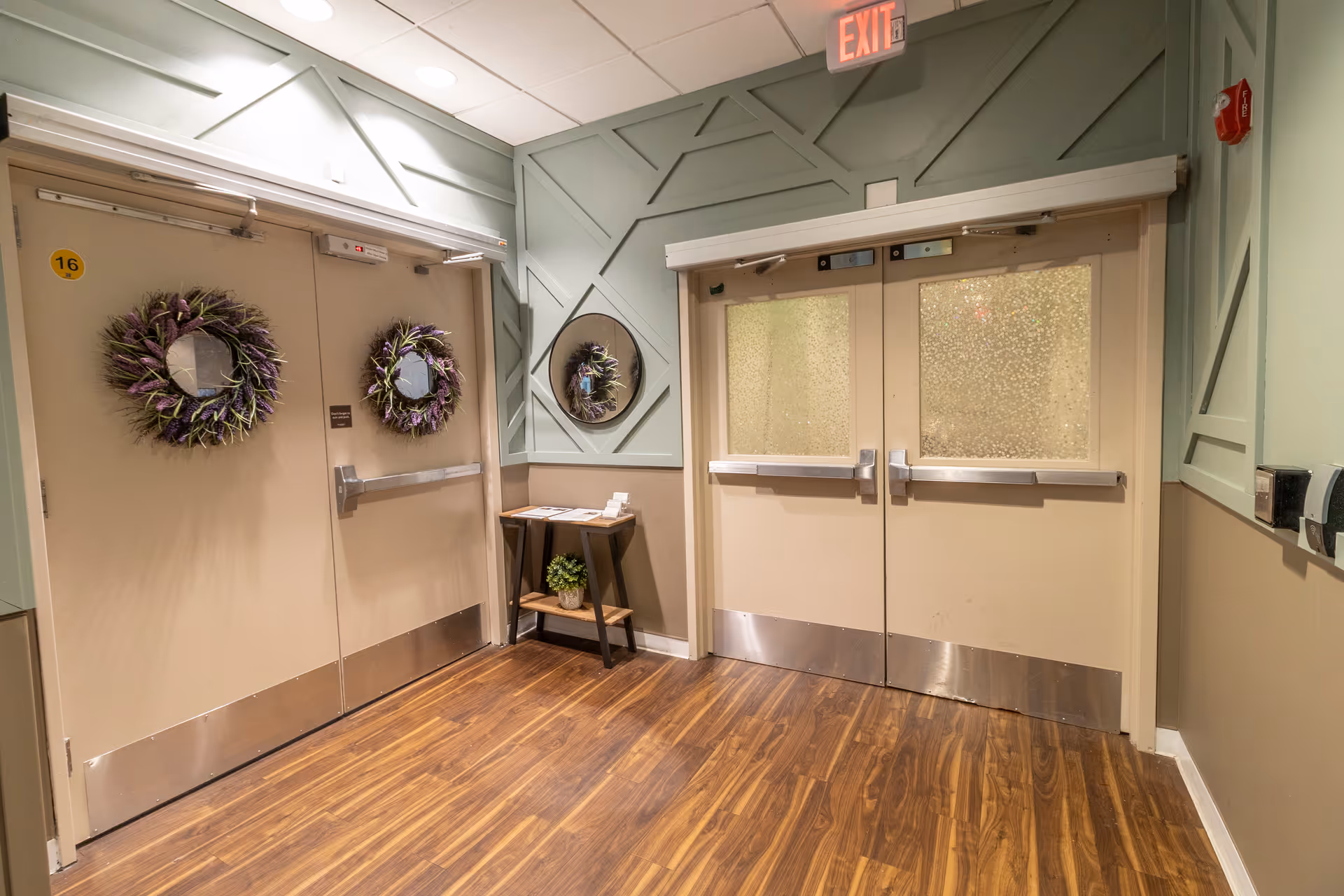 Interior hallway with two sets of beige double doors (one set decorated with wreaths), a small table with decor, and wood flooring.