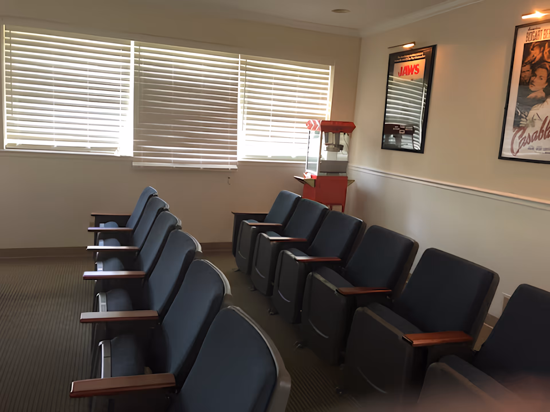 A small theater-style room with two rows of blue cushioned seats with wooden armrests facing a blank wall. There are three windows with white blinds partially closed on the left wall. On the right wall, there are framed movie posters for 'Jaws' and 'Casablanca'. A red popcorn machine is positioned in the corner near the posters.