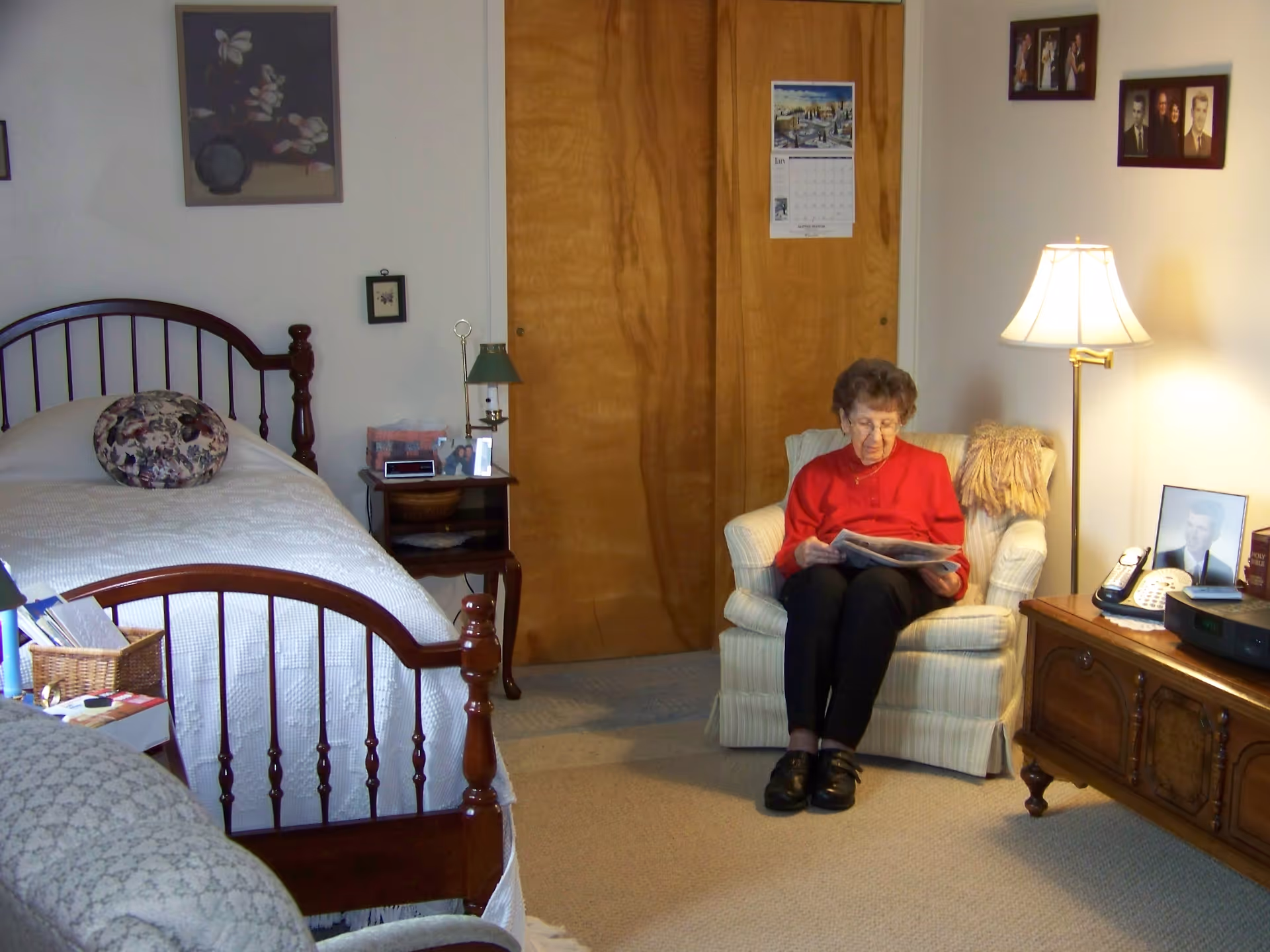 A woman sits reading in a cozy bedroom with a bed, armchair, floor lamp, and wooden furniture.
