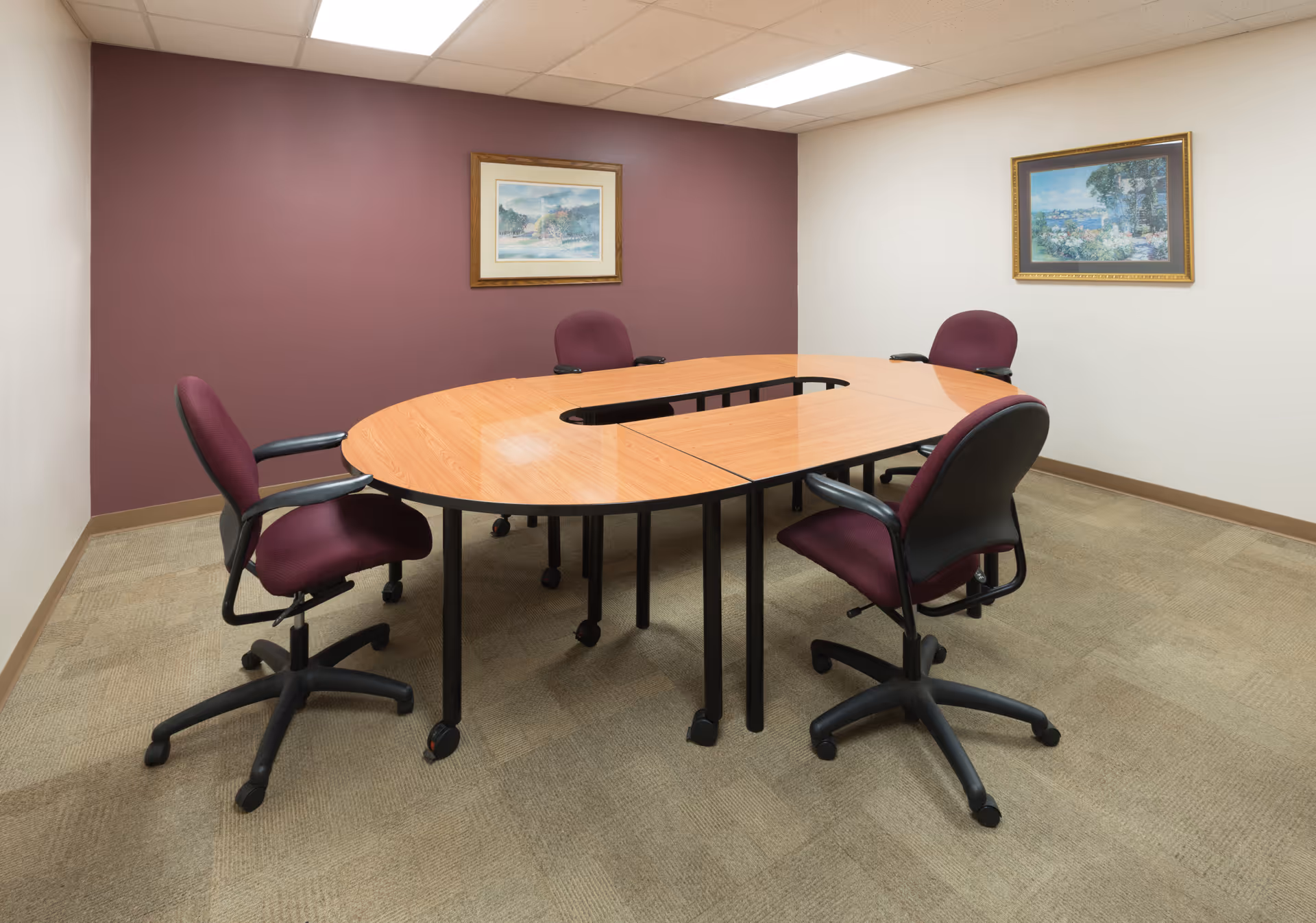 A small conference room with a U-shaped wooden table surrounded by four maroon office chairs on wheels. The room has beige carpet, a white ceiling with fluorescent lights, and two framed landscape paintings on the walls, one on a maroon accent wall and the other on a white wall.
