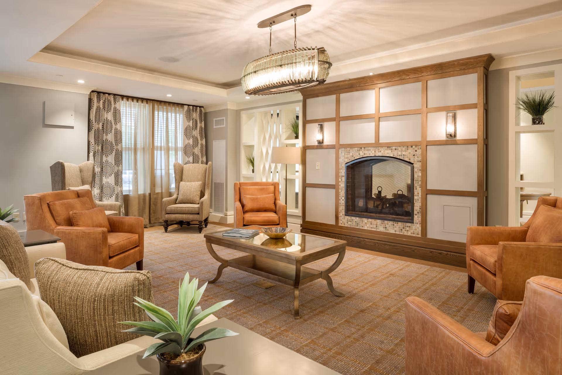 A cozy living room with a large fireplace framed by wood and tile, surrounded by several armchairs in tan and brown tones. A glass-top coffee table sits in the center on a patterned carpet. The room is softly lit with a modern chandelier and recessed ceiling lights. Decorative plants and curtains with a circular pattern adorn the space.