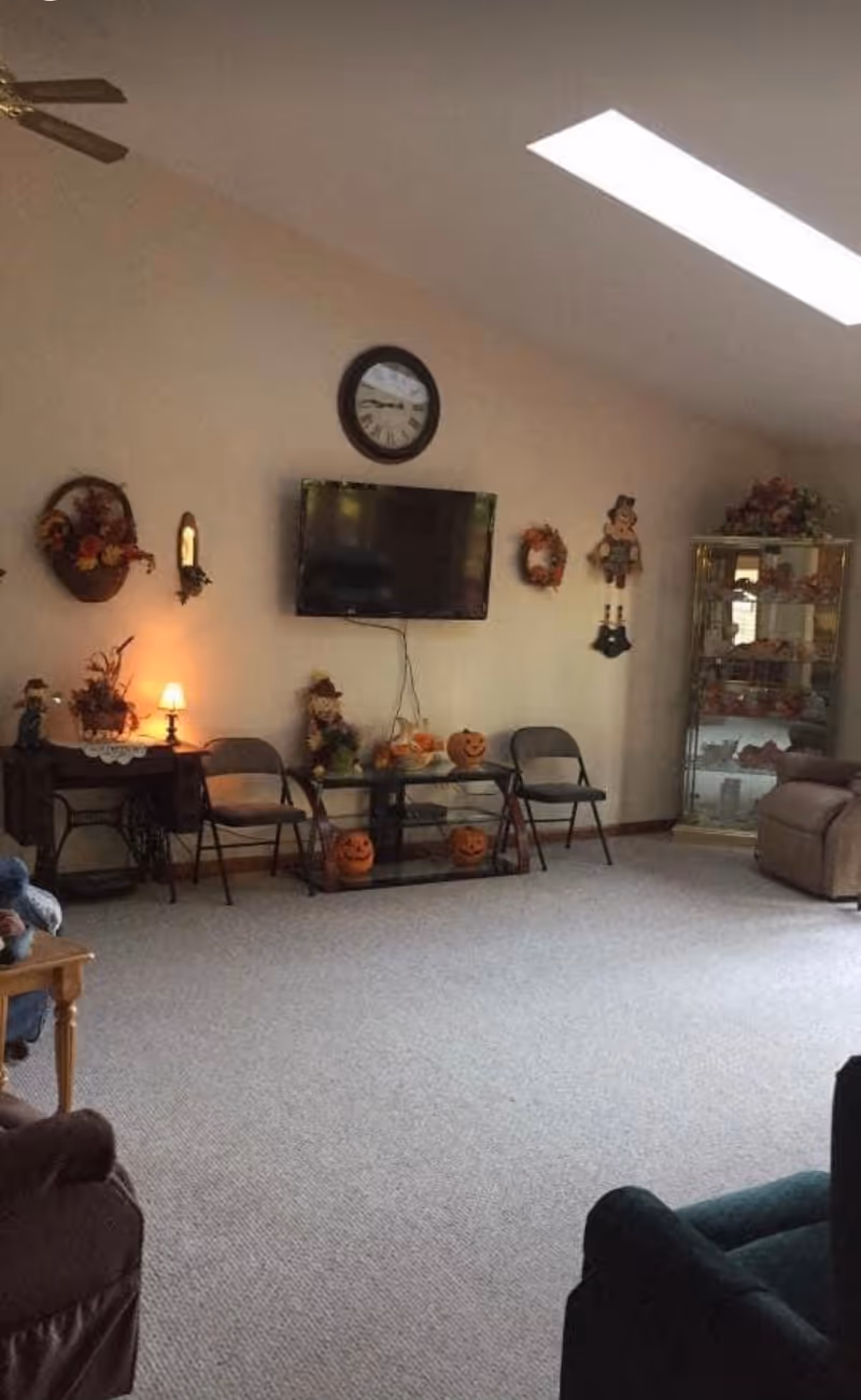 A cozy living room with a beige carpet and slanted ceiling featuring a skylight. The room has a wall-mounted TV beneath a round clock, flanked by autumn-themed decorations including wreaths and pumpkin ornaments. There are two folding chairs and a glass table with more pumpkin decorations. A small table with a lamp and decorative items is on the left, and a glass display cabinet with various items is on the right. Comfortable recliner chairs are partially visible in the foreground.