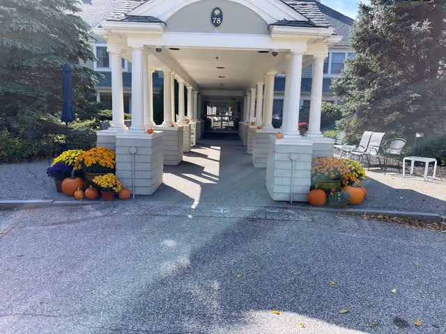 Covered entrance walkway of a building with white columns and a peaked roof displaying the number 78. The entrance is decorated with orange pumpkins and pots of yellow and purple flowers on both sides. There are chairs and a small table on the right side near a large tree.