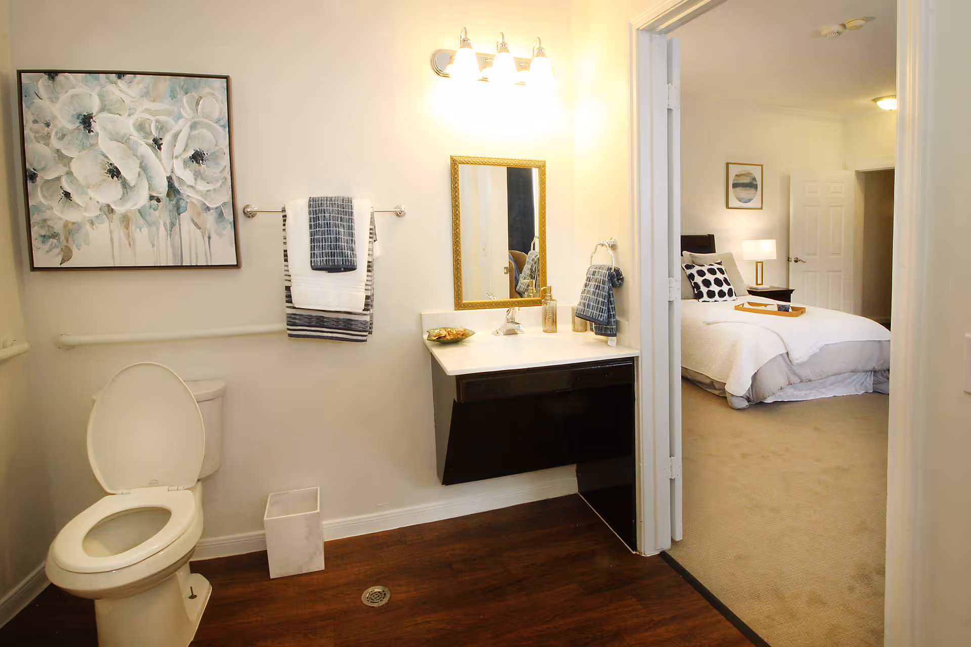Well-lit bathroom with a toilet, wall-mounted vanity and towels, opening into a bedroom.