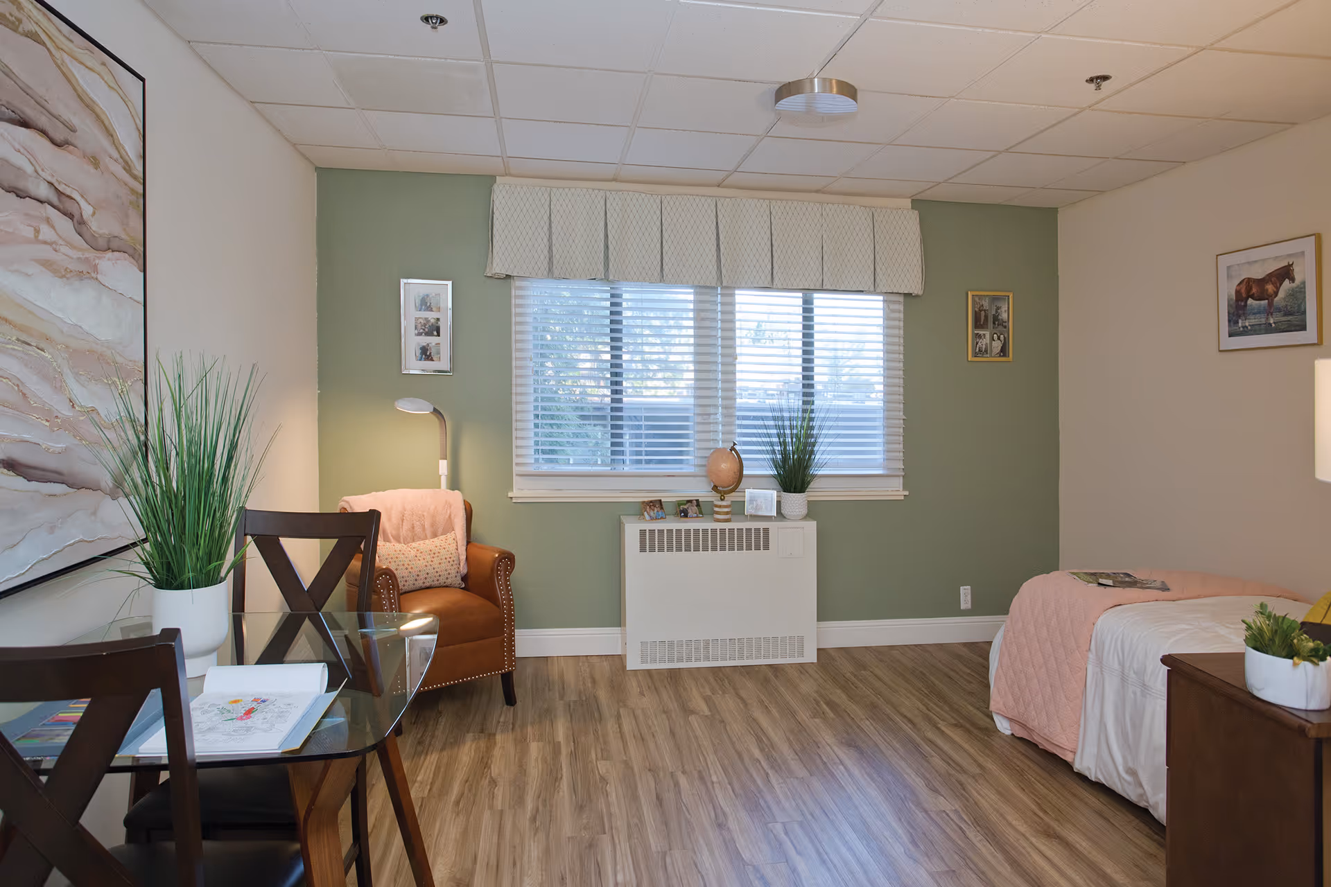 A cozy and well-lit bedroom in a senior living facility featuring a single bed with a pink quilt, a wooden nightstand with a small plant, a brown leather armchair with a pink throw and pillow, a glass-top table with two chairs, and a large window with white blinds and a valance. The walls are painted green and white, decorated with framed pictures and artwork. The floor has wood-style vinyl flooring.