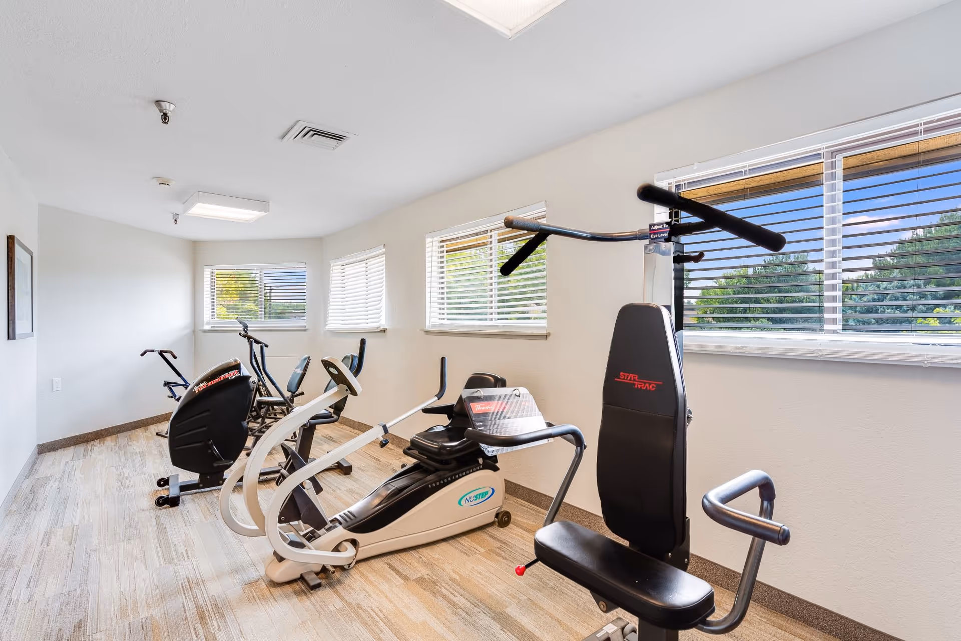 Small indoor fitness room with several exercise machines and windows with blinds.