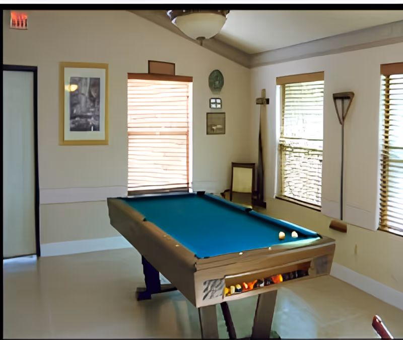 A room with a pool table in the center, featuring a blue felt surface and a few billiard balls on it. The room has three windows with blinds partially closed, a framed picture on the wall, and some decorative items including a wooden paddle and a chair in the corner. The walls are light-colored and the floor is smooth and light-toned.